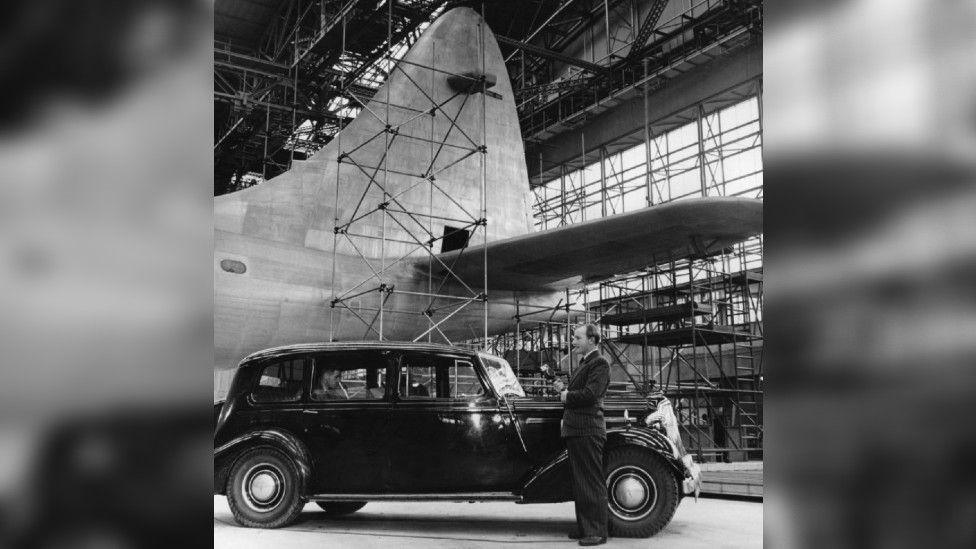 A black and white photo of a BBC reporter in front of the partially-completed Brabazon inside a large aircraft hangar