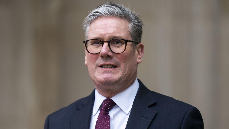 A man with grey hair and black-rimmed glasses photographed from the shoulders up. He is wearing a dark suit, white shirt and purple tie. 