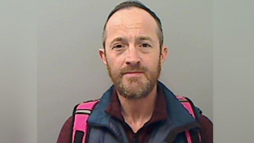 A mugshot of the convicted man who has a slightly ginger beard closely cropped - receding hairline and is wearing a purple shirt with the straps of a pink backpack visible.