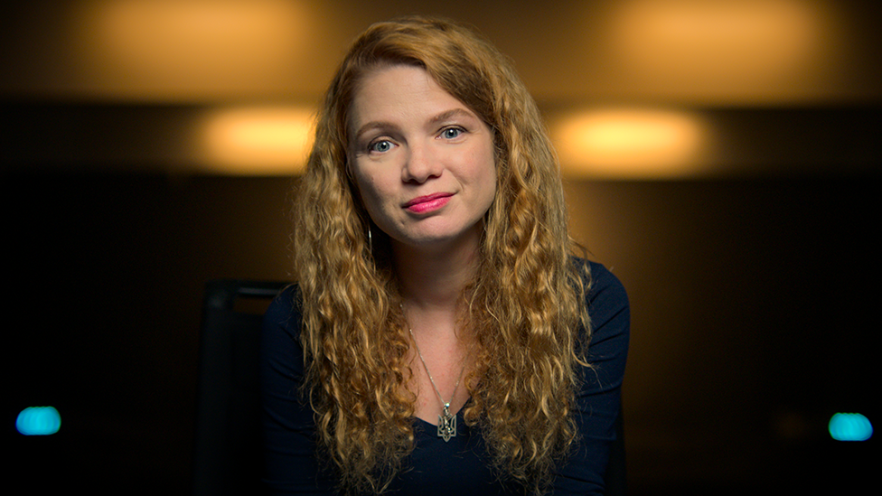 A woman sits against a dimly lit amber background, in an interview-type set up. She's smiling slightly, her long, curly red hair cascading down her shoulders.