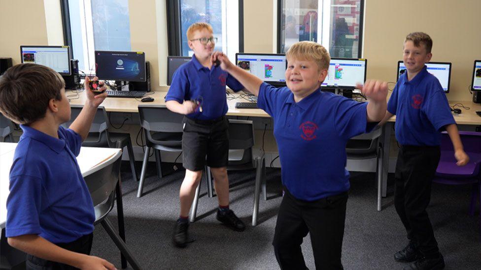 Four Year 6 boys wearing bright blue polo shirts and black trousers and shorts. They are dancing around in a classroom. Behind them is a long row of tables, each with a computer monitor and keyboard and in front of them chairs. 