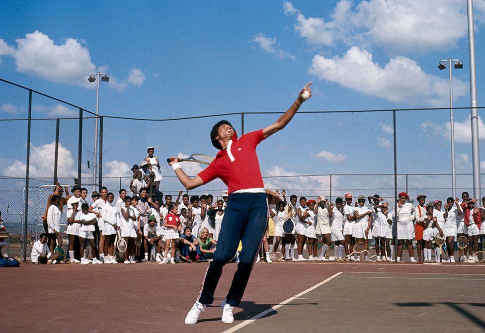 Arthur Ashe in red shirt and navy blue tracksuit bottoms serves as a crowd of South African children in tennis whites watch him from behind a tennis court fence in Soweto - November 1973.