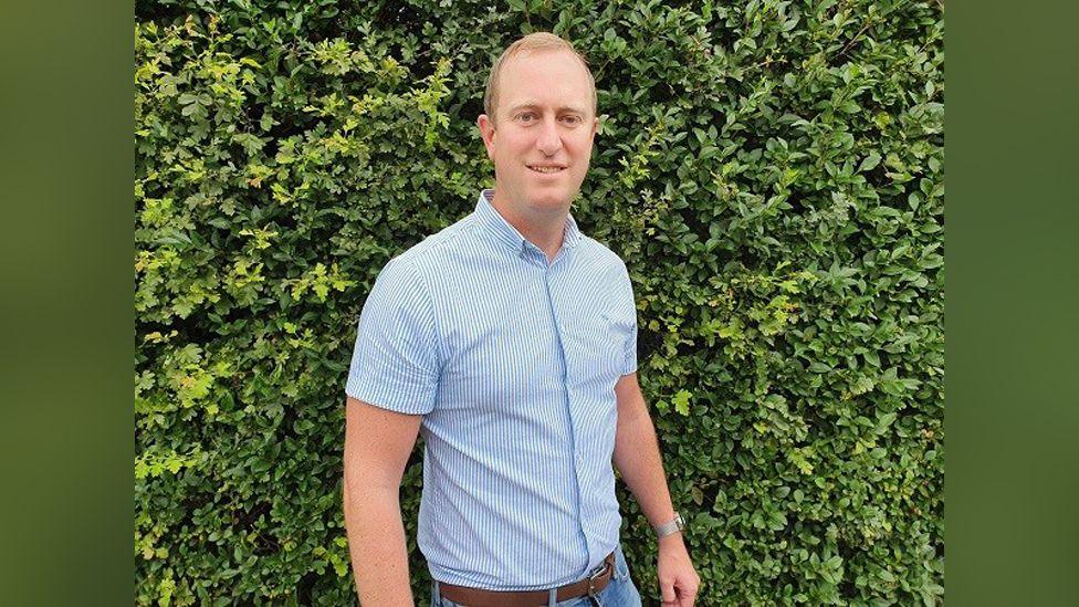 Man with white and blue striped shirt standing in front of a hedge