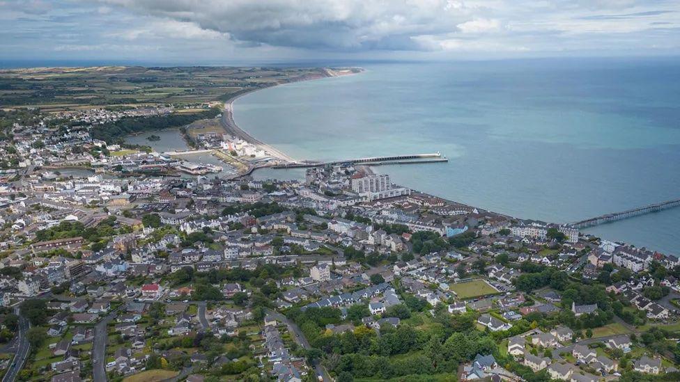 An ariel view of Ramsey, including houses along the arching coastline with the pier going out into the sea.