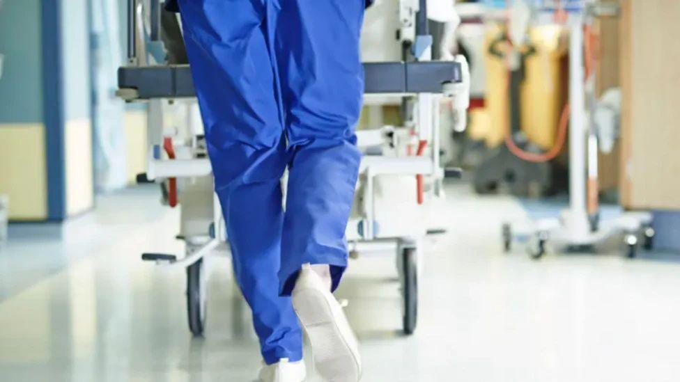 Generic image of a medic wearing blue work trousers pushing a medical trolley along a hospital corridor. The medic's top half is not shown.