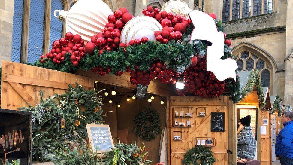 A wooden shed stall with wreaths tied to one of the open doors, and cards displayed on the other. On the roof of the shed there is a large decorative display of baubles, tinsel, and frosty fir leaves.