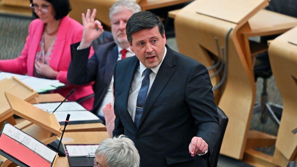 Jamie Hepburn, wearing a dark suit with blue tie, points to his left while speaking in the Scottish Parliament chamber 