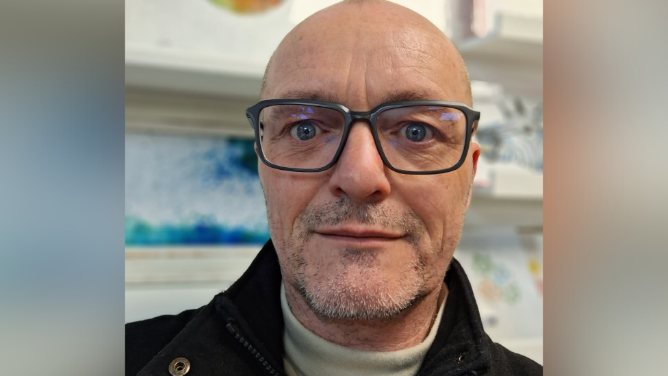 Paul Dumper wearing glasses and a dark jumper pictured in front of paintings in his art studio