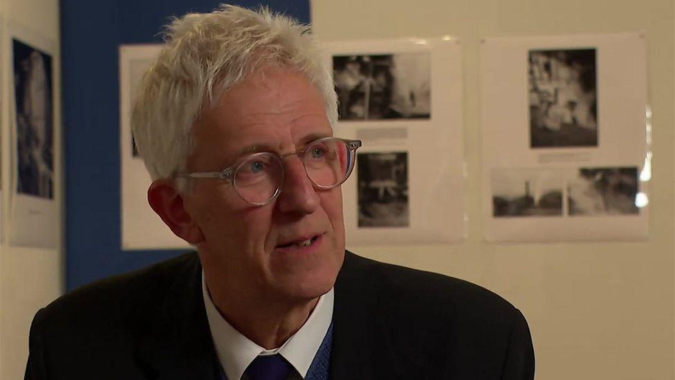 Daniel pictured at the exhibition in London. He has short white hair and is wearing glasses and a dark suit. Images from the exhibition can be seen in the background