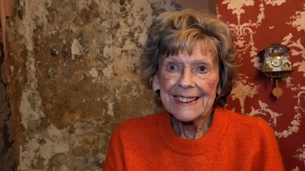 Margaret Chappell, 93, stands wearing a bright orange jumper with immaculate hair and make up standing in front of a wall completely covered in mould