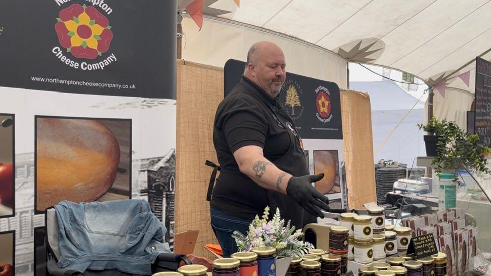 A bald man with tattoos, black apron and rubber gloves is stood gesturing to a wide variety of food products, such as condiments, meat and cheese. He is stood in front of a black sign with reads 'Northampton Cheese Company' with large close ups of food on the sign. He is in a market tent, with bunting in the corner.