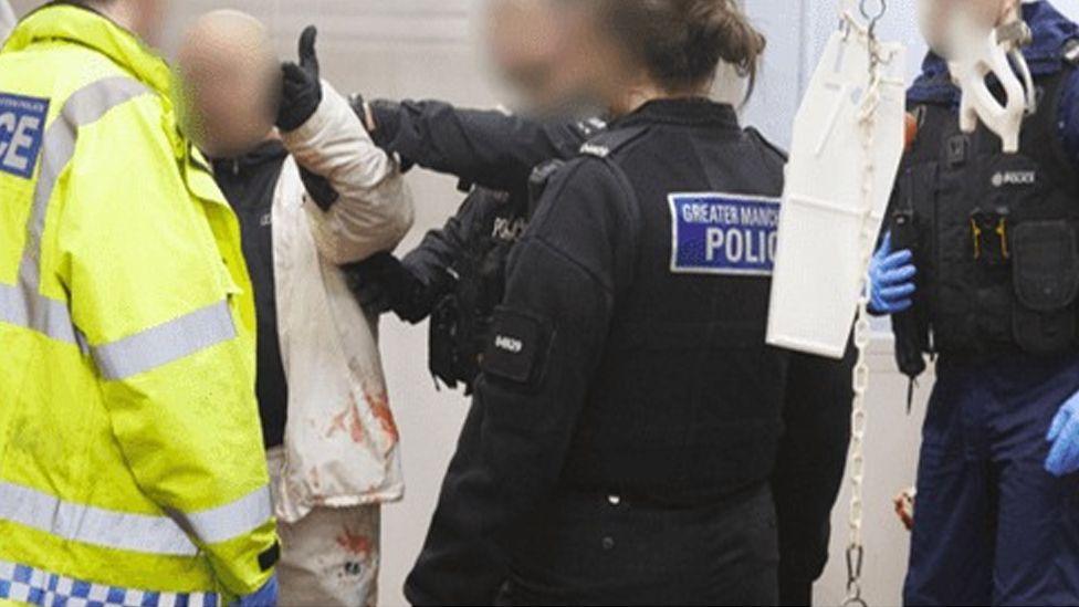 Four police officers in uniform, one in a hi-vis jacket at the abattoir. They are talking to a man in white scrubs whose face is blurred.