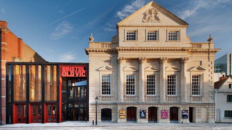 The frontage of the Bristol Old Vic theatre. It is a large historic sand coloured building with tall sash windows and large pillars. On the left there is a more modern addition to the building, with black and red metal beams and a large sign saying 'Bristol Old Vic'. 