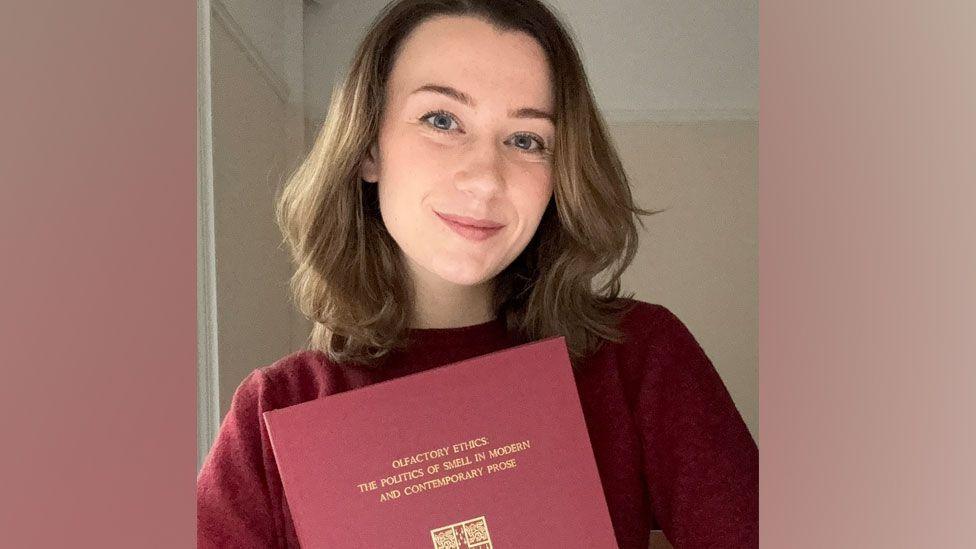 Dr Ally Louks is holding her thesis - a red A4 sized hardback book. Its title is written in gold lettering on the front. Dr Louks is smiling at the camera. She has shoulder length light brown hair, blue eyes and is wearing a burgundy jumper. 