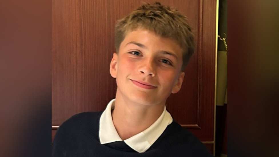 Joshua Travis, a 14-year-old boy with mousey brown hair. He is wearing a school uniform and appears to be standing in front of a wardrobe and he his smiling.