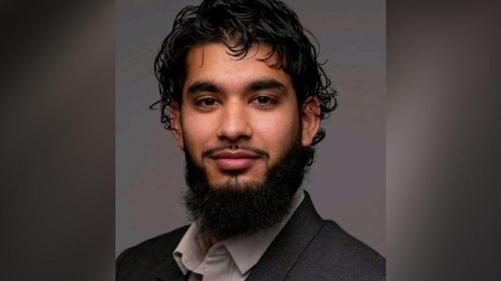 Adam Khan, a man who is sitting looking at the camera and smiling. He has black hair and black facial hair. He is wearing a grey suit jacket and light grey shirt.