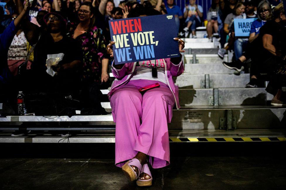 An woman in a pink suit, sitting in a crowd, holds a sign which reads WHEN WE VOTE, WE WIN