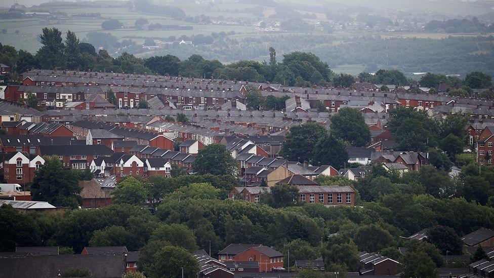Rows of terraced houses and rooftops in Blackburn, there are a lot of trees to the front and the moors in the distance on an overcast day. 