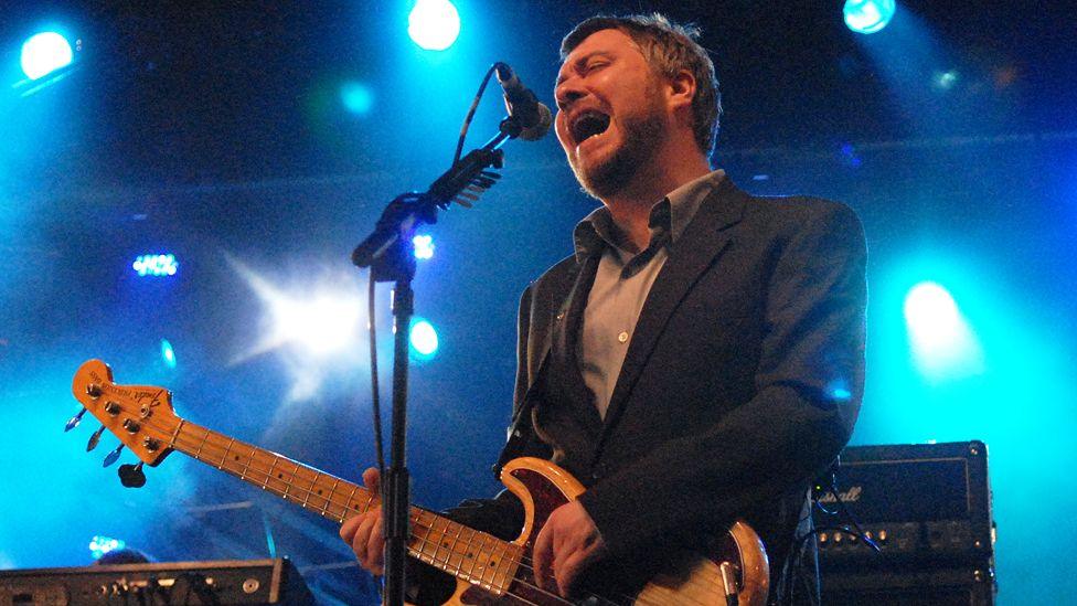 Singer Jimi Goodwin, with short hair and a beard and wearing  black suit jacket and grey shirt, plays a guitar left-handed in front of black speakers and bright blue lights