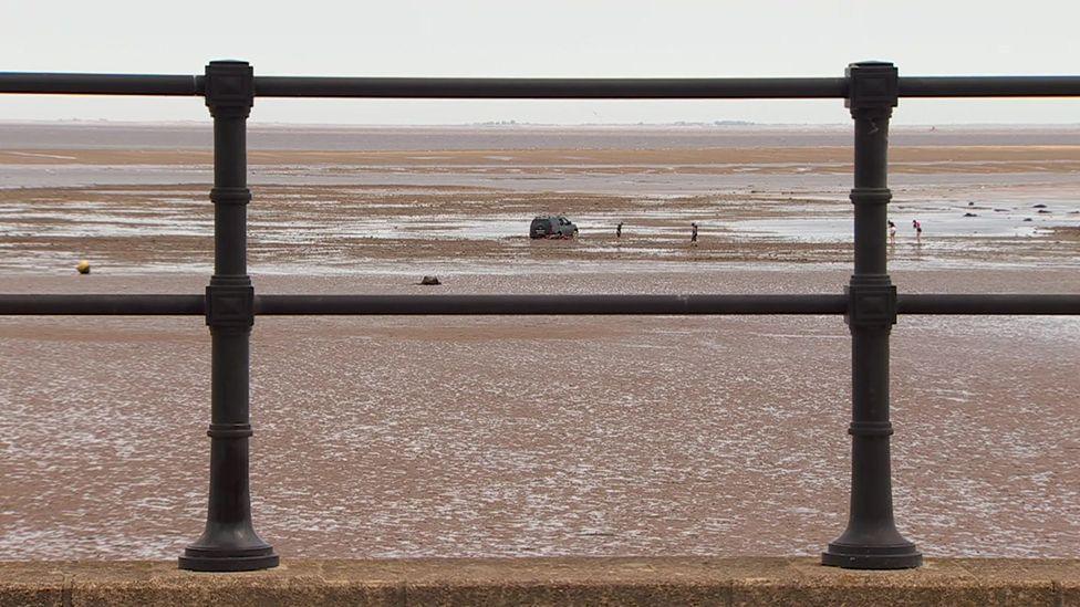 A view of the stranded car in the distance on the beach through black iron railings