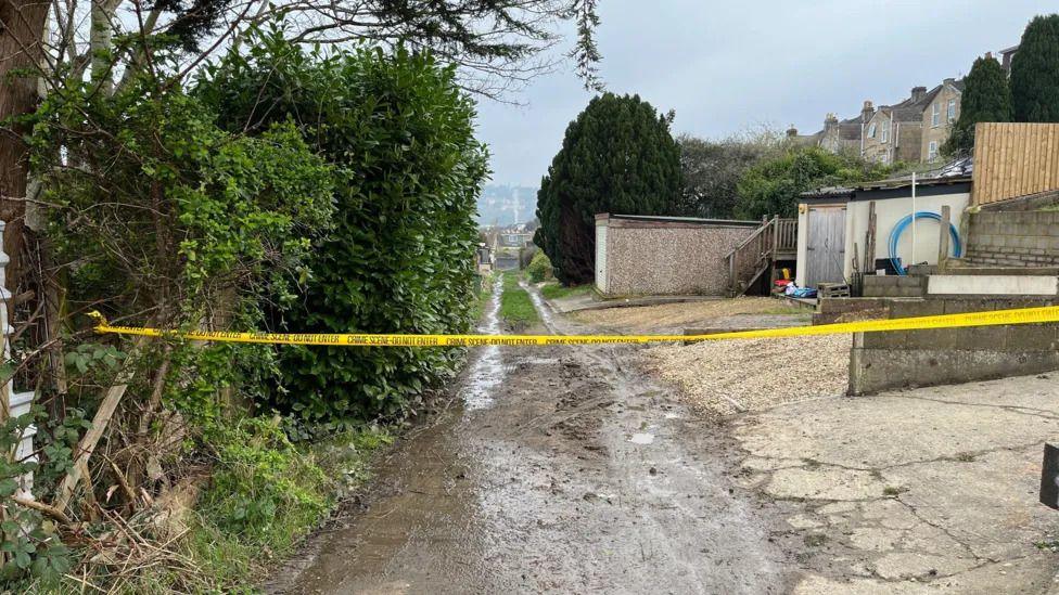 Police tape stretching across a muddy footbath in Bath, with trees and outbuildings visible