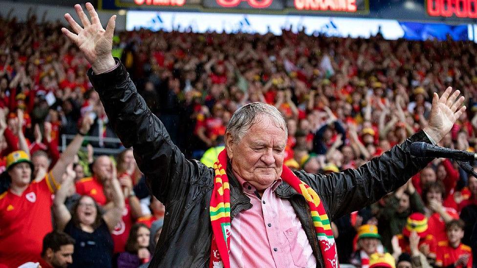 Dafydd Iwan in Cardiff City stadium. He has short grey hair and is wearing a light pinky-red shirt, with a leather jacket open over it and a Wales football scarf in red, green and yellow. There is a microphone to his right and he has his hands raised in the air while looking down. Behind him is a stand full of fans, many in red Wales football shirts and/or wearing red, green and yellow bucket hats which have become associated with the Wales team. At the top a digital display reads Cymru 0 - 0 Ukraine.
