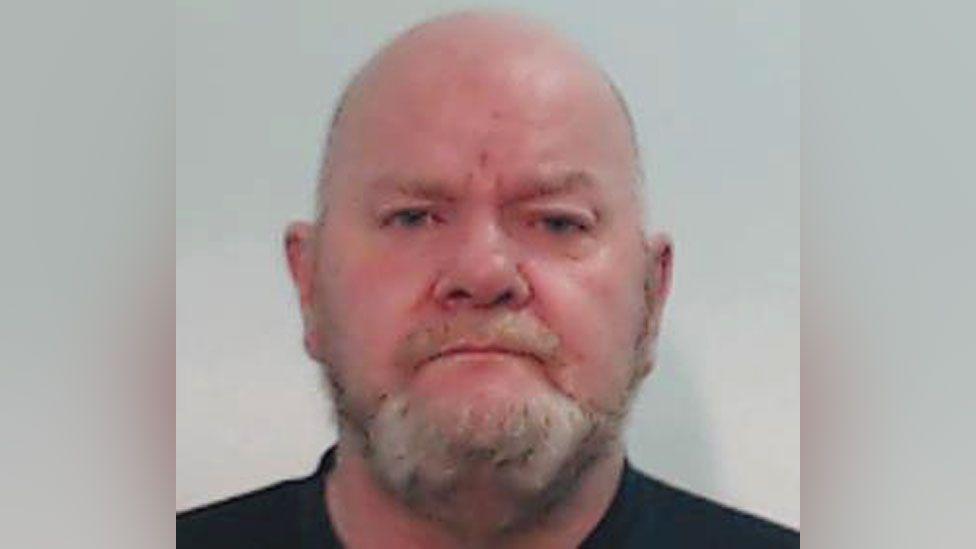 A man in his 50s with a bald head and a beard in a police mugshot