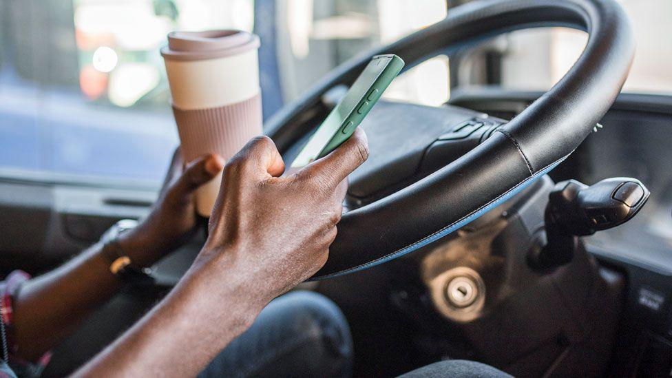 A man sits in a stationary vehicle in front of a steering wheel holding a cup of coffee in one hand and a phone in the other