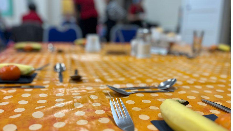 A table covered in an orange wipeable tablecloth with white dots. It is laid for meal with knives, forks and spoons, as well as salt and pepper. At each laying is a banana and tomato. People can be glimpsed milling around in the background.