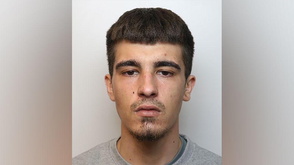 Police mugshot of Kurtis Knee with a shaved moustache and goatee beard, dark hair and dark eyes, wearing a grey top