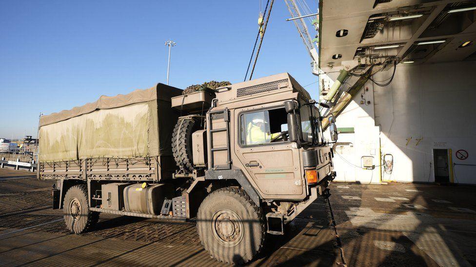 Army truck with person seen driving, moving into the cargo area of a large ship.