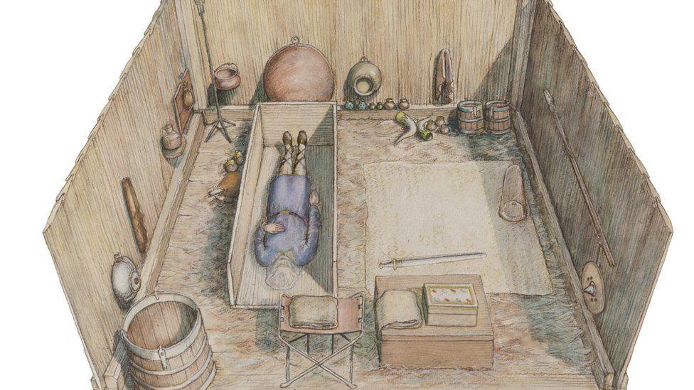 An artist's impression of the tomb at Prittlewell. It shows a wooden clad rectangular pit. On the left is a wooden coffin with a man's body wearing a blue tunic within. Behind its head is a camping stool. To its right is a box. Behind it and to the right of the coffin is a rug with a sword lying upon it. Hanging off the wooden walls are pots, a shield and a spear. There are drinking horns and apples on the ground, plus wooden barrels. 