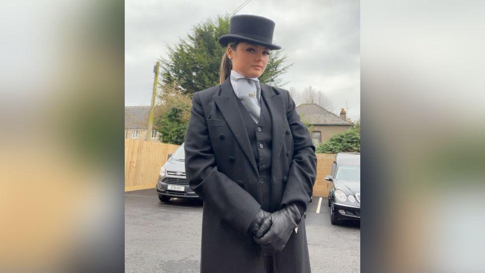 A young woman stood in black suit and black top hat on with black hearses in the background