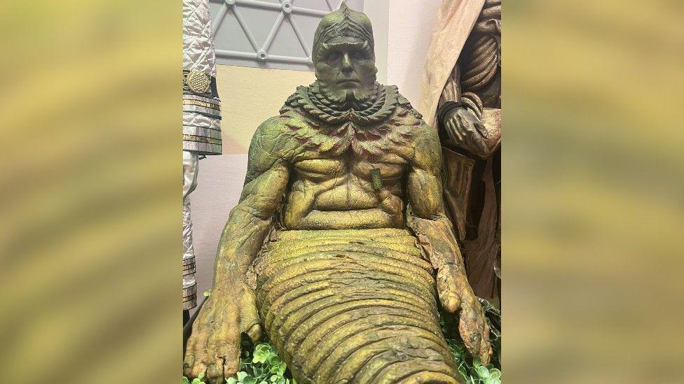 Lord Kiv, who is a green character from Doctor Who, is sat in the middle of the image. It a slug-liked character that has long arms and rippled skin.