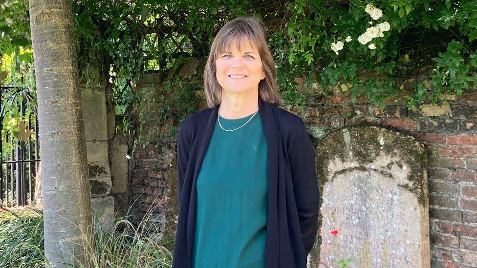 Sarah Smith wearing a green dress and navy cardigan, standing next to a tree