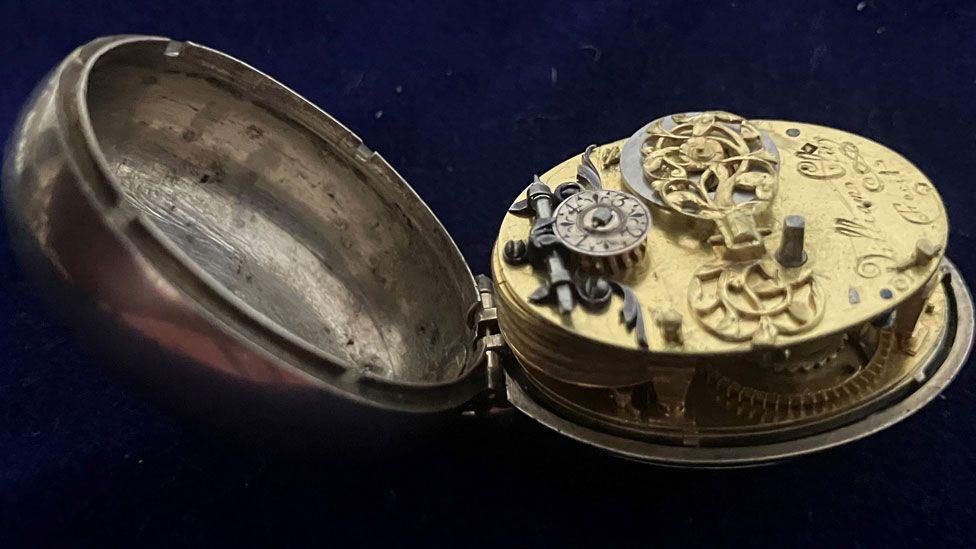 A 17th Century silver pocket watch, opened up so the domed case is on the left and the golden and silver coloured cogs and wheels of the interior are on the right, against a black background