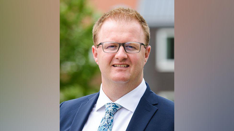 The headteacher of Esher High School, Andy King, pictured with the school grounds blurred into the background. Mr King has short ginger hair and is wearing grey glasses and a navy suit with a white shirt. 