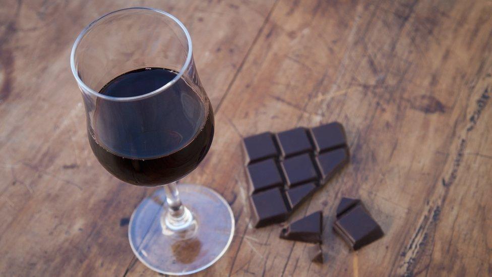 A glass half full of red wine on a table, beside half-eaten dark chocolate.