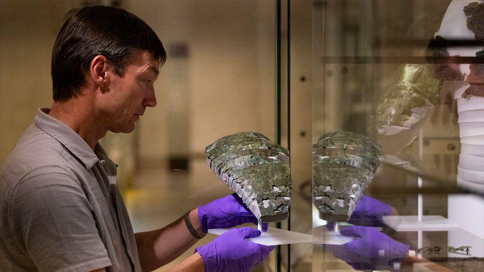 A man wearing purple gloves delicately places a Roman artefact into a glass display unit with some reflection of his hands and the armour