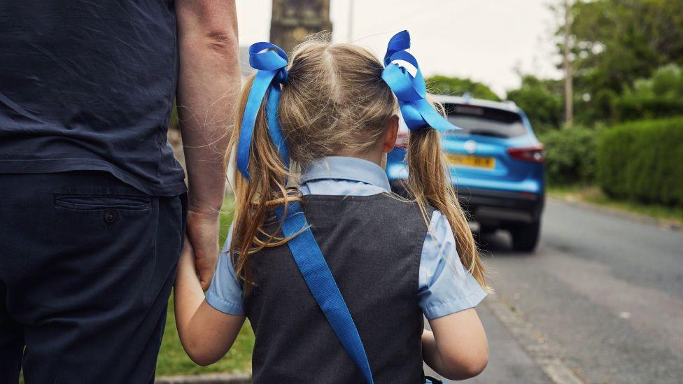 A young girl, with blue ribbons tied in her hair and wearing a blue shoulder bag, walks down the street holding the hand of a man. A blue car is in the background