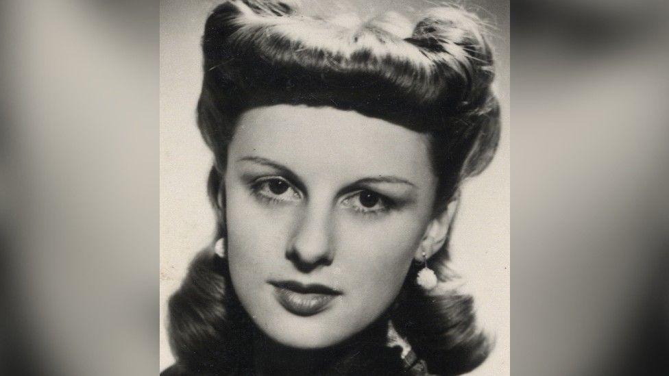 A black and white headshot image of Carol in her twenties looking down the lens of the camera. She is wearing drop white ball earrings and her hair is highly styled.