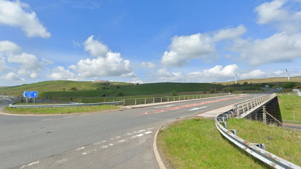 Junction 37. It is a two-lane road with a blue sign which reads 'The South M6'. There are cars in the distance and two wind turbines on the hills to the right.