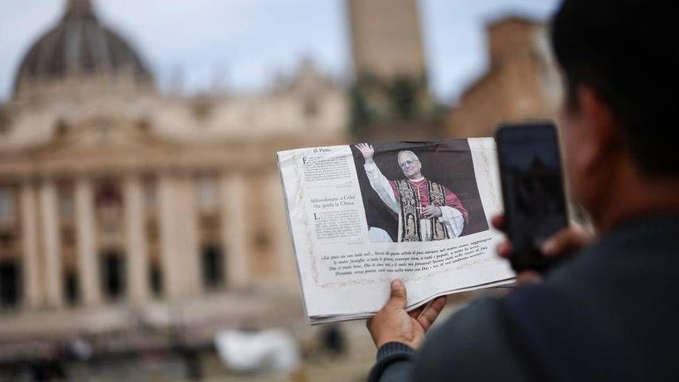 A person takes a picture of a newspaper with the picture of the Pope on the front page in front of the Sistine Chapel