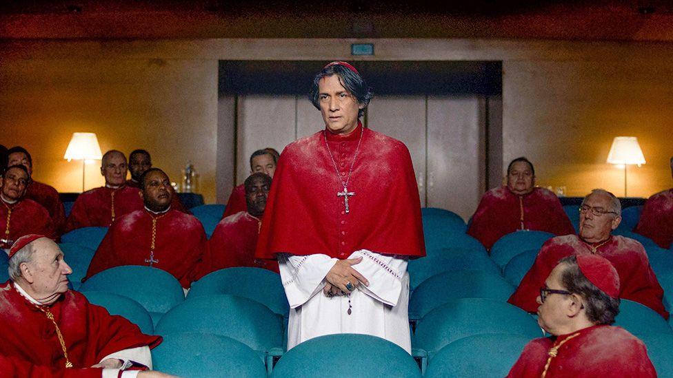 A group of cardinals dressed in red robes and white garments are seated in a room with blue chairs. One central figure, Cardinal Benitez, standing and wearing a cross necklace, is positioned prominently in the middle of the image. The background features wooden paneling and two lit lamps on either side.