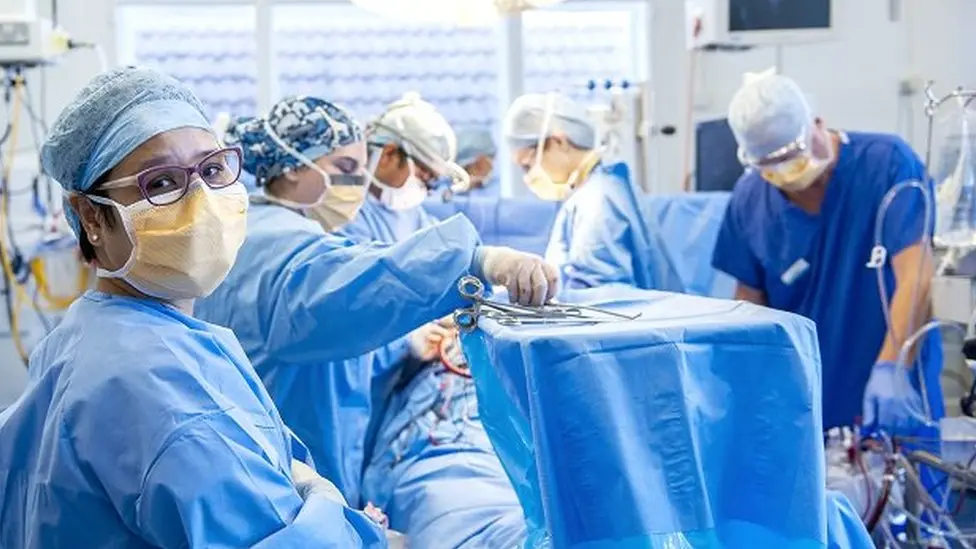 A hospital theatre where surgery is being carried out. Five surgeons gather around an unseen patient, who is covered with blue paper cloths. The surgeons all wear matching blue gowns and wear masks and caps. On a high tray are surgical instruments, with one surgeon reaching for a scalpel.
