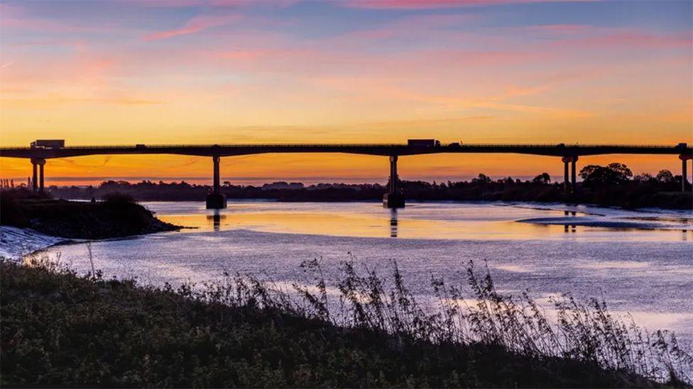 A long shot of the Ouse Bride silhouetted in the sunset with lorries driving across