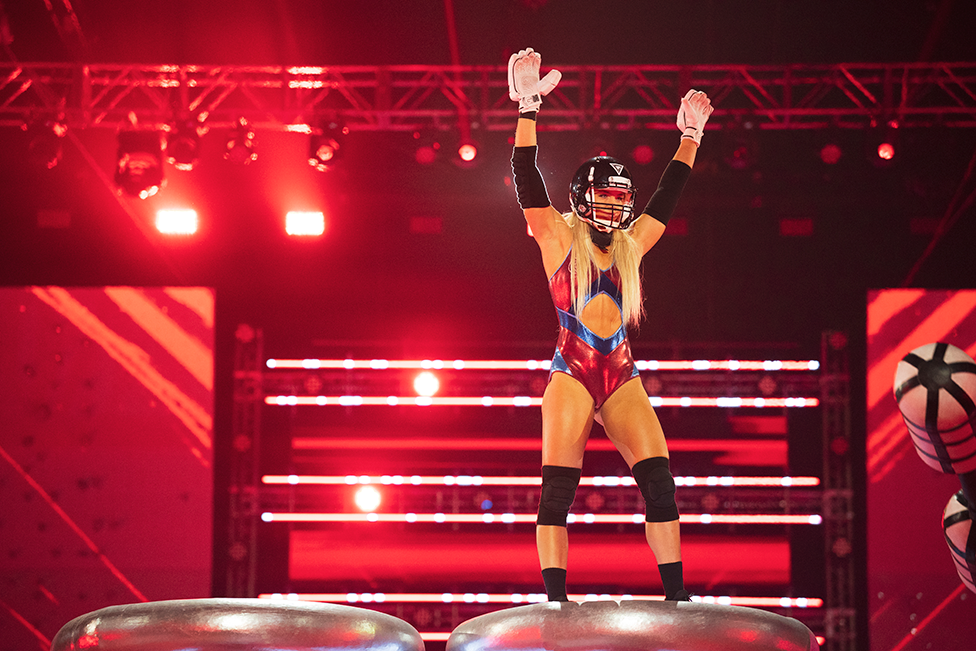 A woman with long blonde hair, wearing a black helmet and a red and blue Gladiators outfit, stands on a podium during the show with her arms in the air.