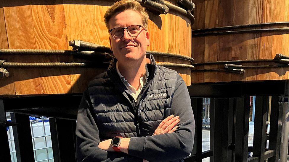 Ben Murphy, who wears glasses, standing with his arms folded in front of two large wooden distillery containers. 