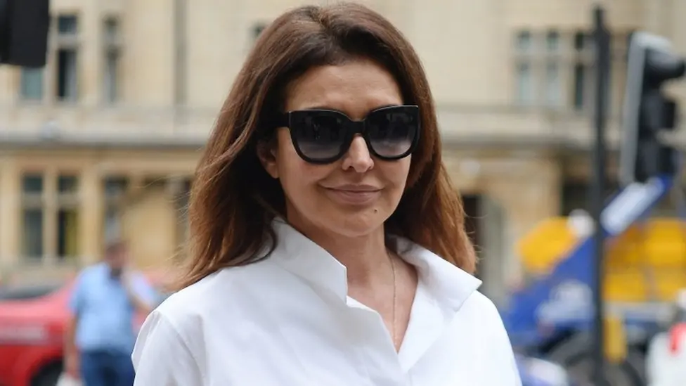 A picture of Zamira Hajiyeva, who has shoulder-length brown hair and is wearing sunglasses, walking along an unknown street.