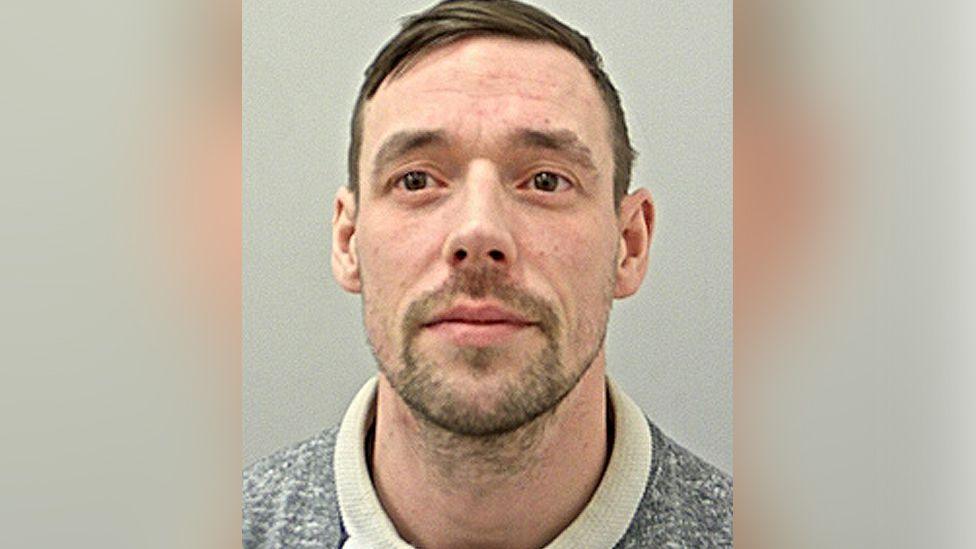 Police custody image of Shaun Fuller who has brown eyes and brow hair with brown stubble. He is wearing a cream coloured grey jumper.